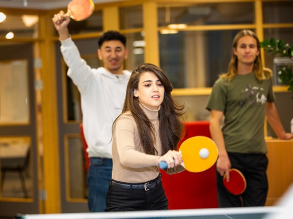 En ung kvinna med långt hår spelar bordtennis. Bilden fångar precis när hon slår på bollen. I bakgrunden står två unga män och väntar på sin tur.