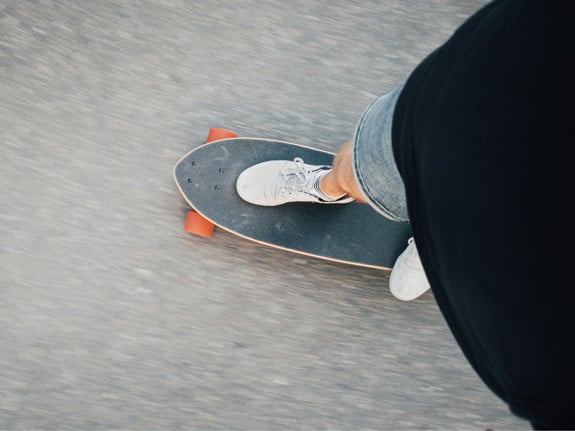 En skateboard sedd uppifrån på asfalt. Man ser fötterna på personen som åker skateboard.