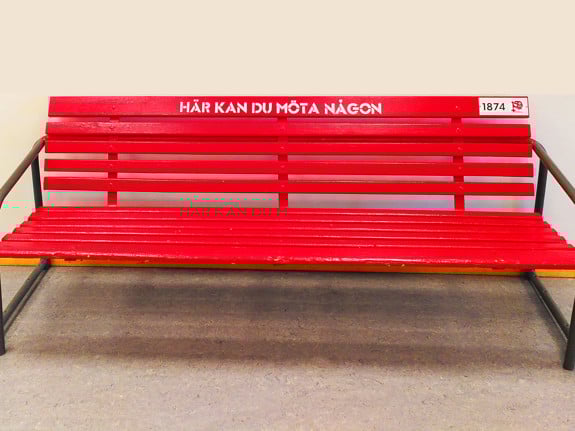 En röd parksoffa i trä. På översta ribban står det med vit text: Här kan du möta någon