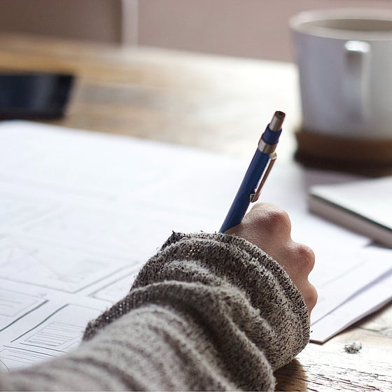 närbild hand som skriver med en bläckpenna i ett stort anteckningsblock. I bakgrunden står en mugg.