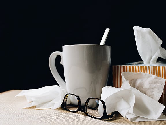 På ett bord står en kaffemugg med en sked som sticker upp. Bredvid står en kartong med näsdukar. Framför muggen och kartongen ligger ett par glasögon och en använd näsduk.